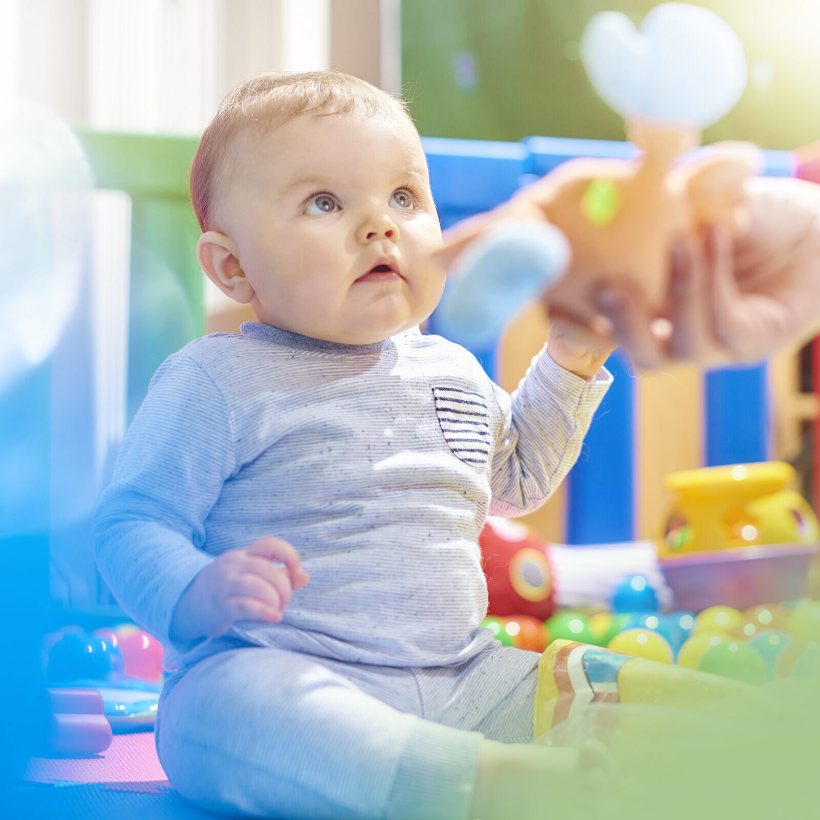 A young baby boy sits playing in his brightly coloured playpen. He discovering how to move and play with his toys. The bright colours, sounds and different textures of the toys are building his sensory perceptions. His mother is passing him one of his cuddly toys.